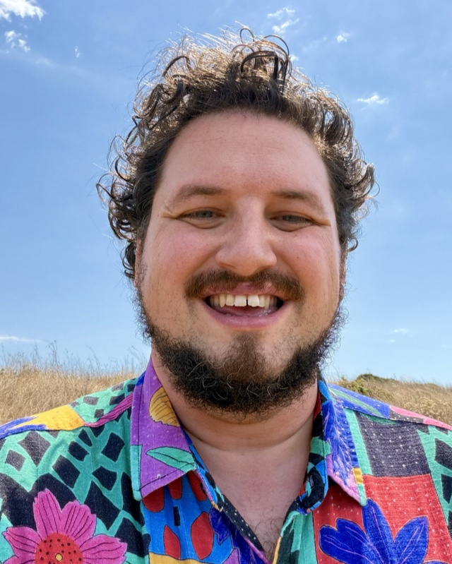 Patrick Angle, smiling, in a collage-print shirt against a blue sky and dry summer grass.